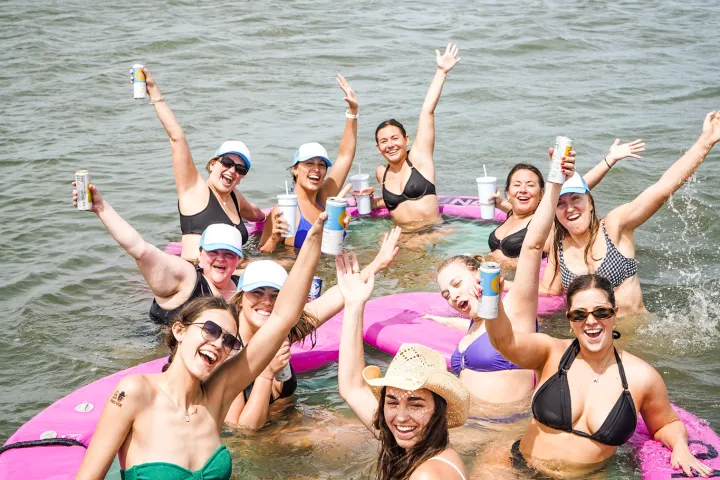 Group of people in swimsuits smiling and waving from a pink circular float in a lake.