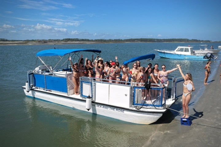 Group of people on a pontoon boat in swimwear near a sandy shore.