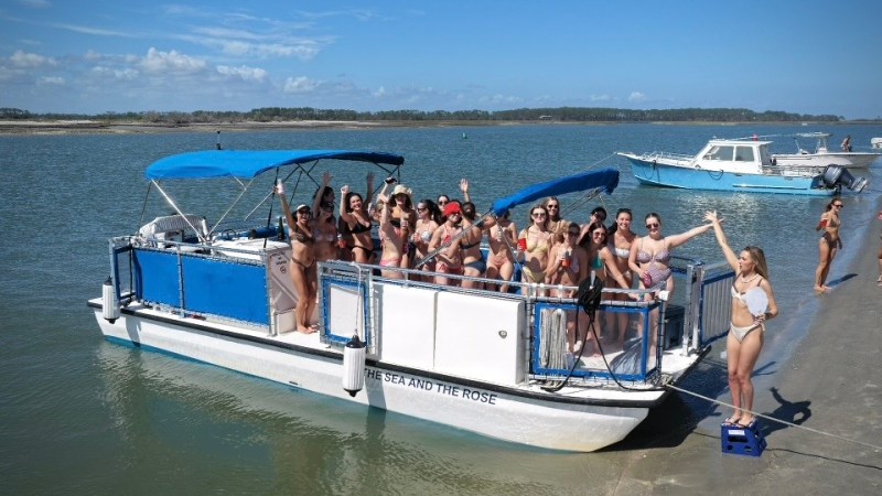 Group of people on a pontoon boat in swimwear near a sandy shore.