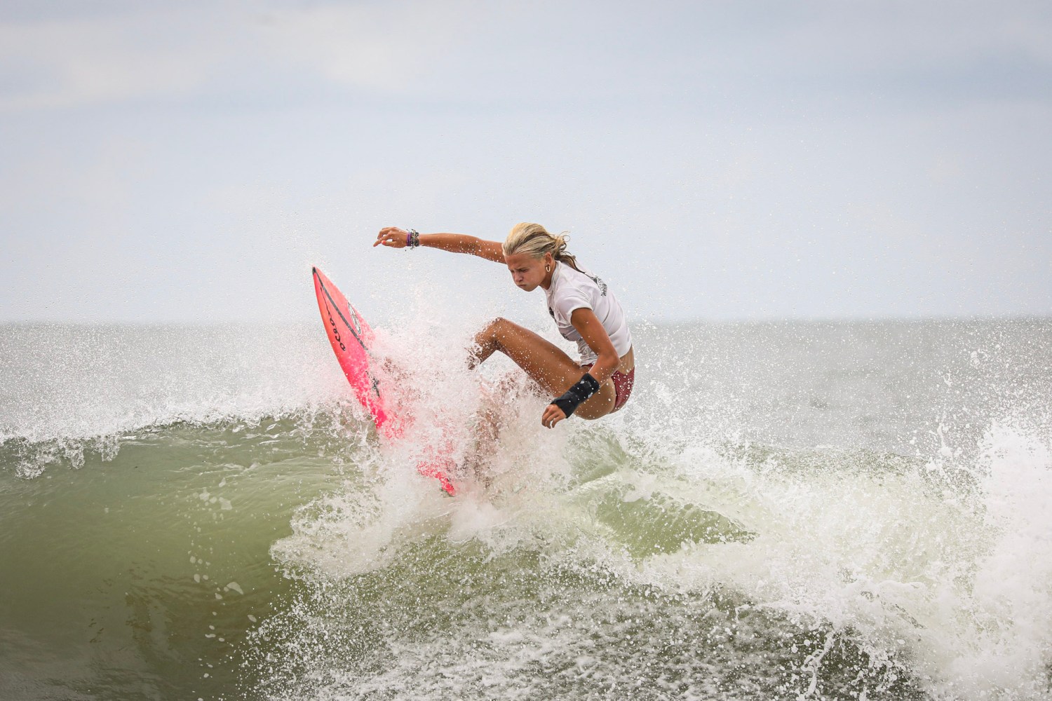Surfer on a pink board riding a wave against a cloudy sky.