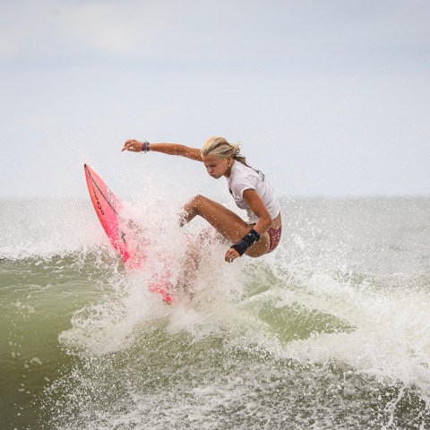 Surfer on a pink board riding a wave against a cloudy sky.