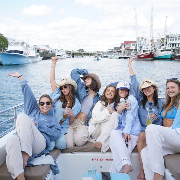 Group of women smiling on a boat, raising glasses, with docks and boats in the background.