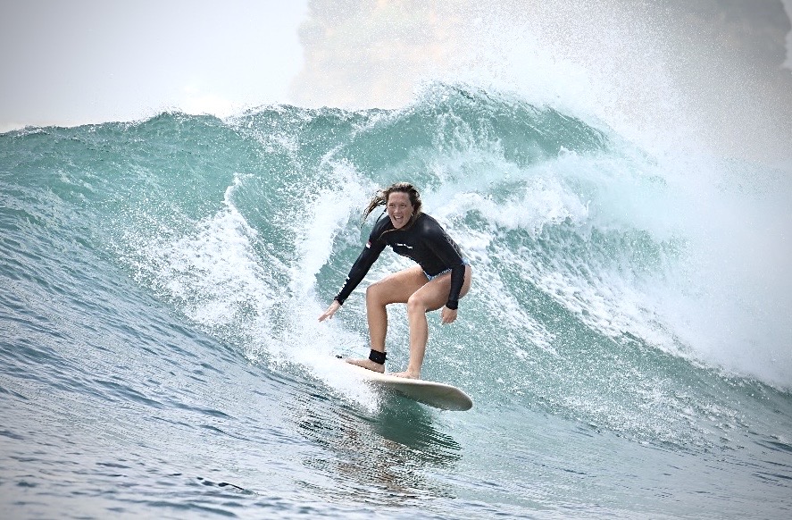 Person surfing on a large wave, wearing a black wetsuit and smiling, with water splashing around.
