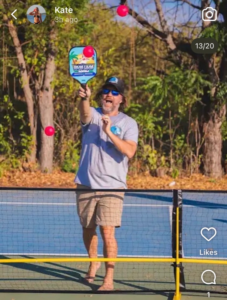 Man on a court juggling pink balls with a paddle in a wooded area.
