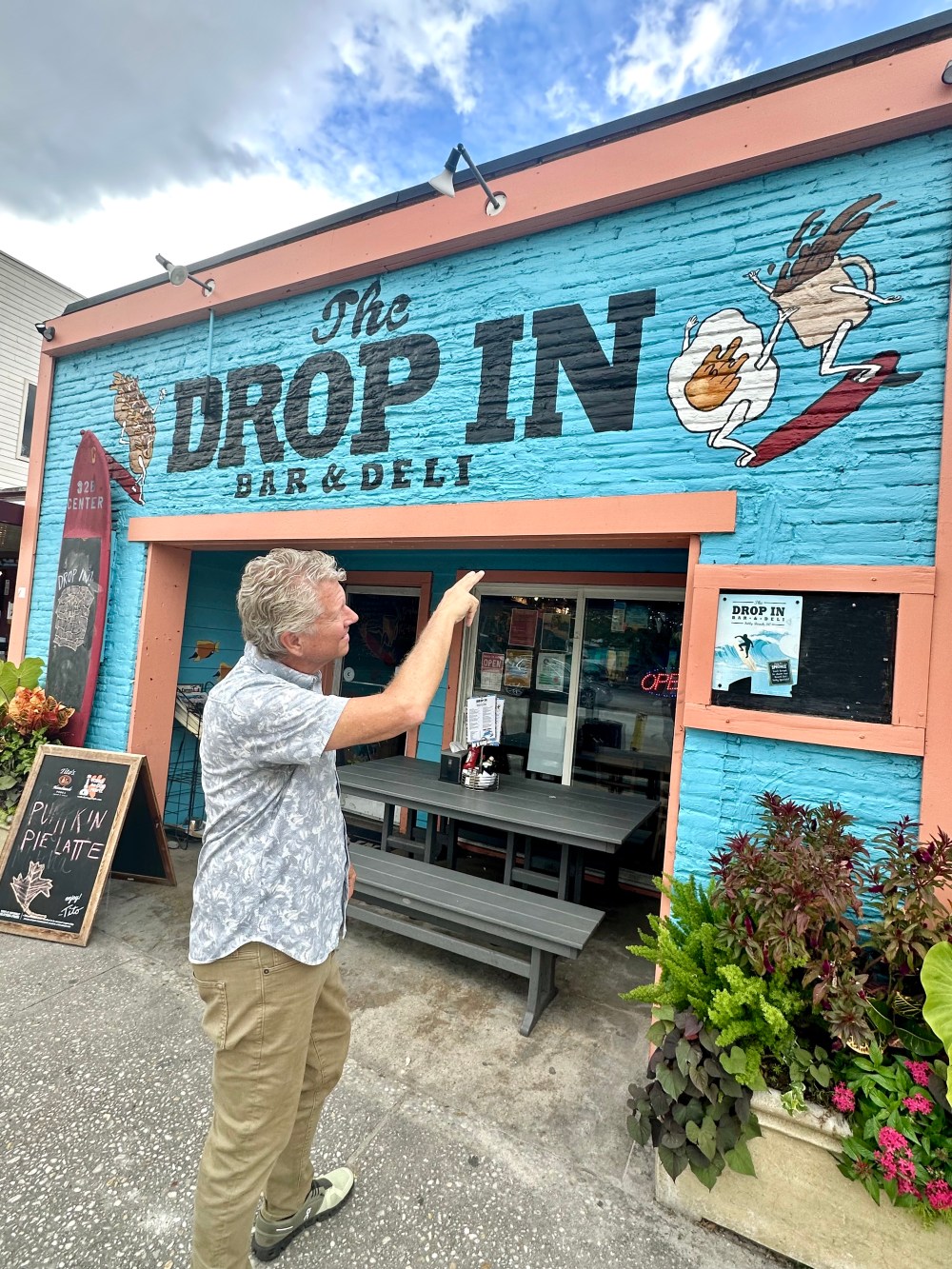 Man pointing at colorful mural of 'The Drop In' bar and deli exterior with plants and bench.