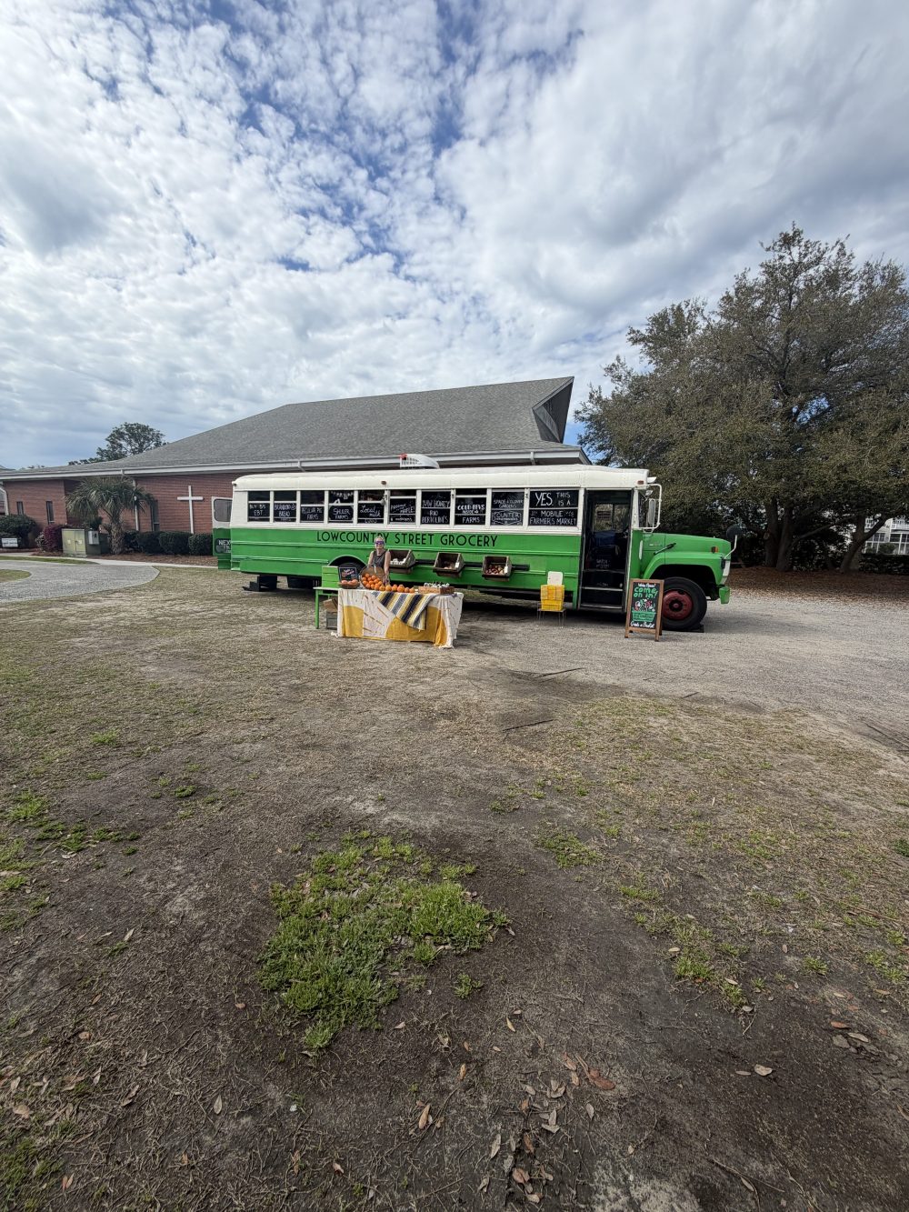 Green grocery bus parked outside a building with trees and a cloudy sky.
