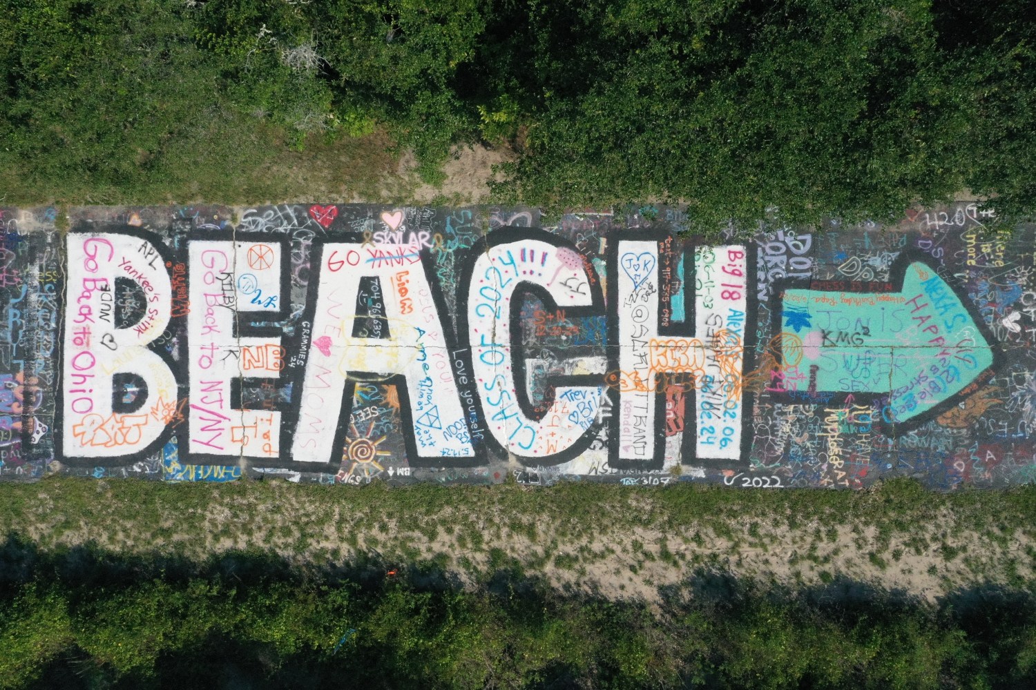 Graffiti art spelling 'BEACH' with colorful designs on a path, surrounded by greenery.